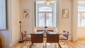 Een moderne eetruimte met een glazen tafel en bruine stoelen, een vloerlamp en een groot raam met uitzicht op gebouwen.