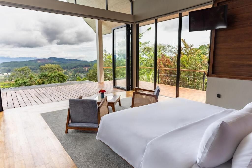 A bedroom with a large bed, a seating area with a table, and a balcony with a view of the landscape.