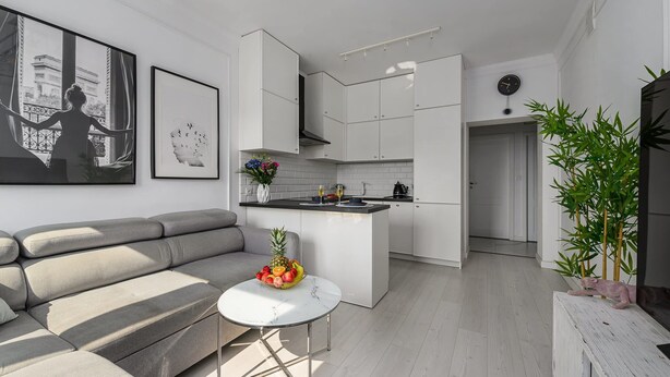 A modern living room with a grey sofa, a small white table with a fruit bowl, and a kitchen area with white cabinets and a black countertop. - Flat With Quiet Courtyard (Warsaw)