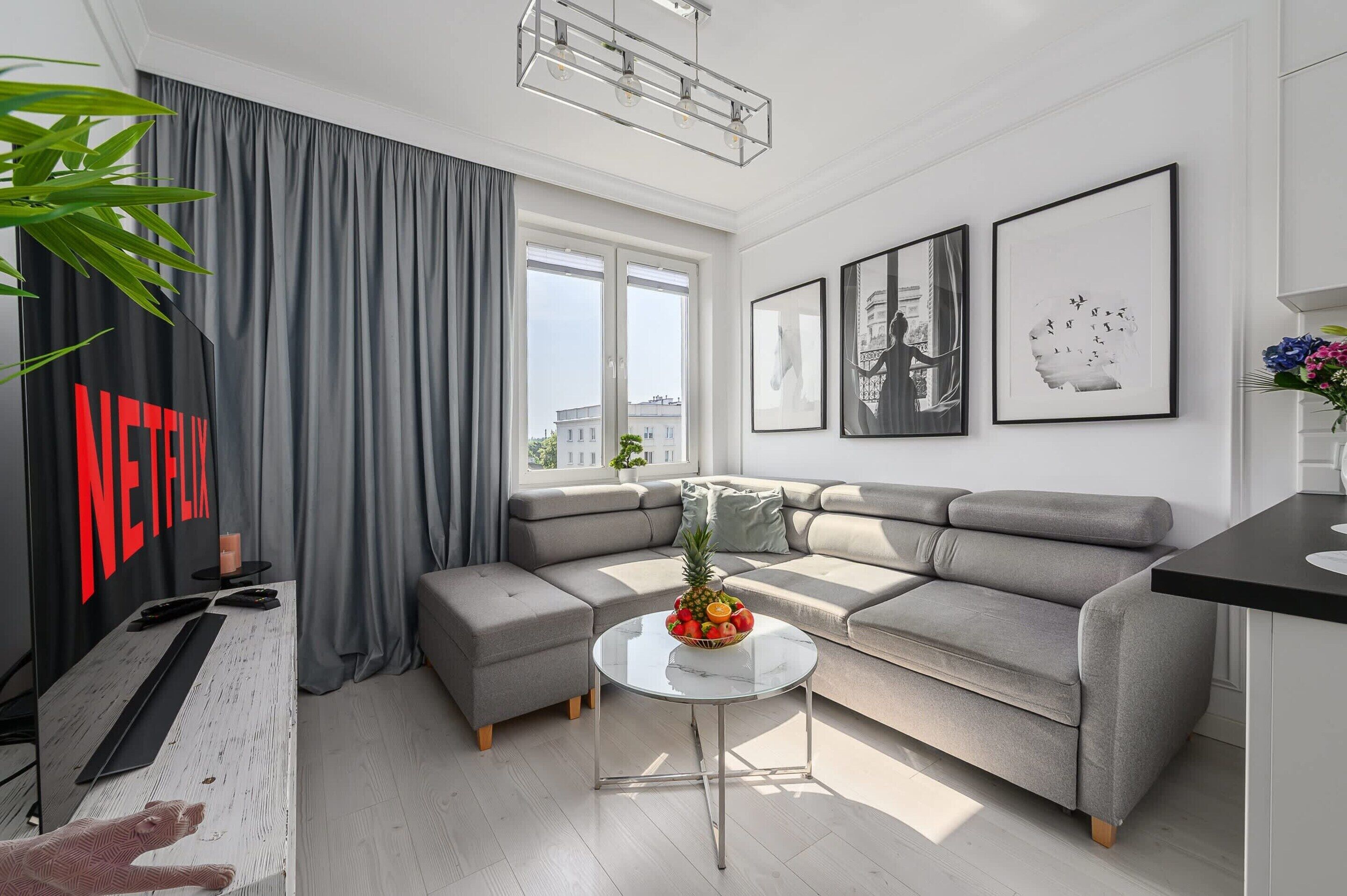 Een moderne woonkamer met een grijze modulaire bank, een ronde salontafel met fruit, en een groot raam met gordijnen.