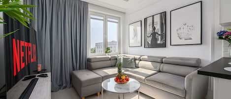 Een moderne woonkamer met een grijze modulaire bank, een ronde salontafel met fruit, en een groot raam met gordijnen.