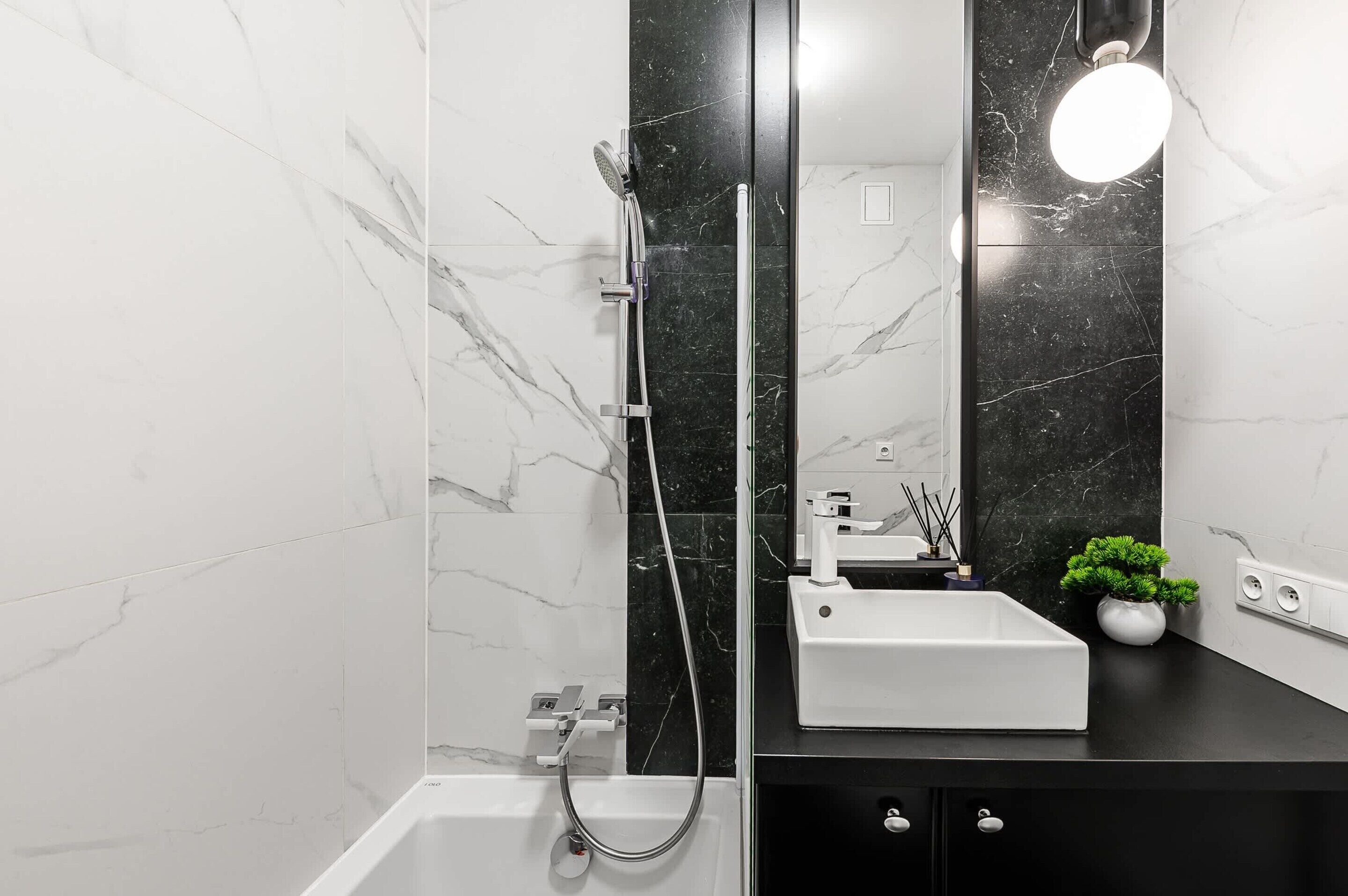 A modern bathroom with a white marble wall, a black marble shower enclosure, a white sink, and a small potted plant.