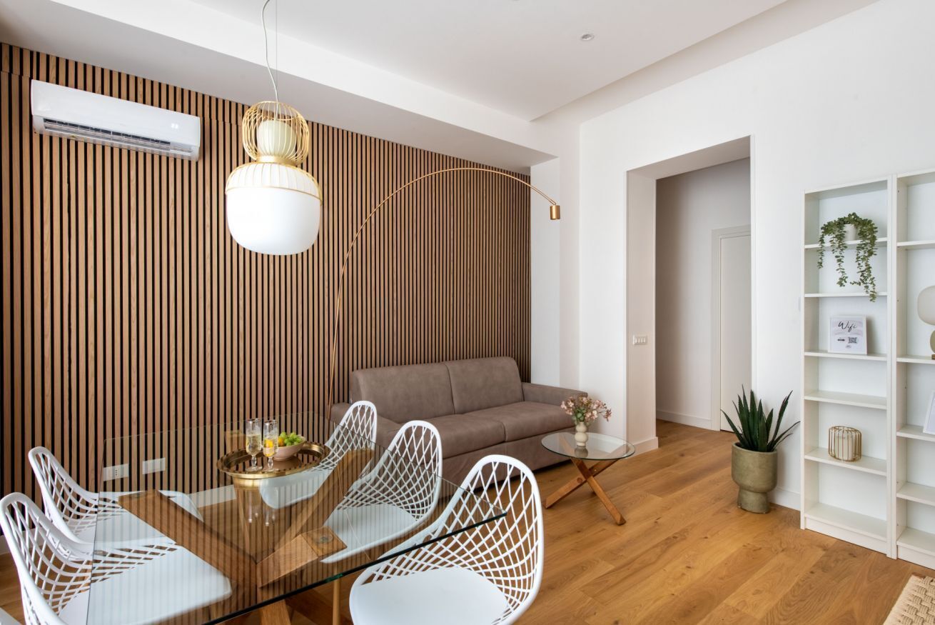 A modern living room with a glass dining table, white chairs, a brown sofa, and a wooden slatted wall.