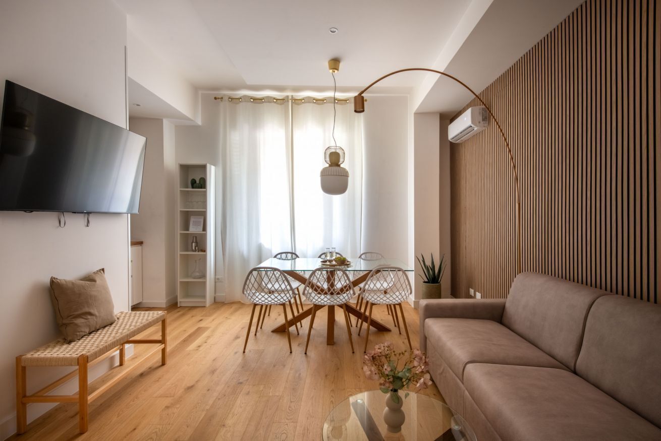 A modern living room with a flat-screen TV, a sofa, a dining table with chairs, and a bench.