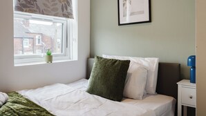 A bedroom with a bed, a window with a view of buildings, a framed picture on the wall, and a bedside table with a lamp. - 4 bed, 4 en-suite house (Warrington)