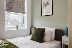 Um quarto com uma cama, uma janela com vista para prédios, um quadro na parede e uma mesa de cabeceira com uma luminária.