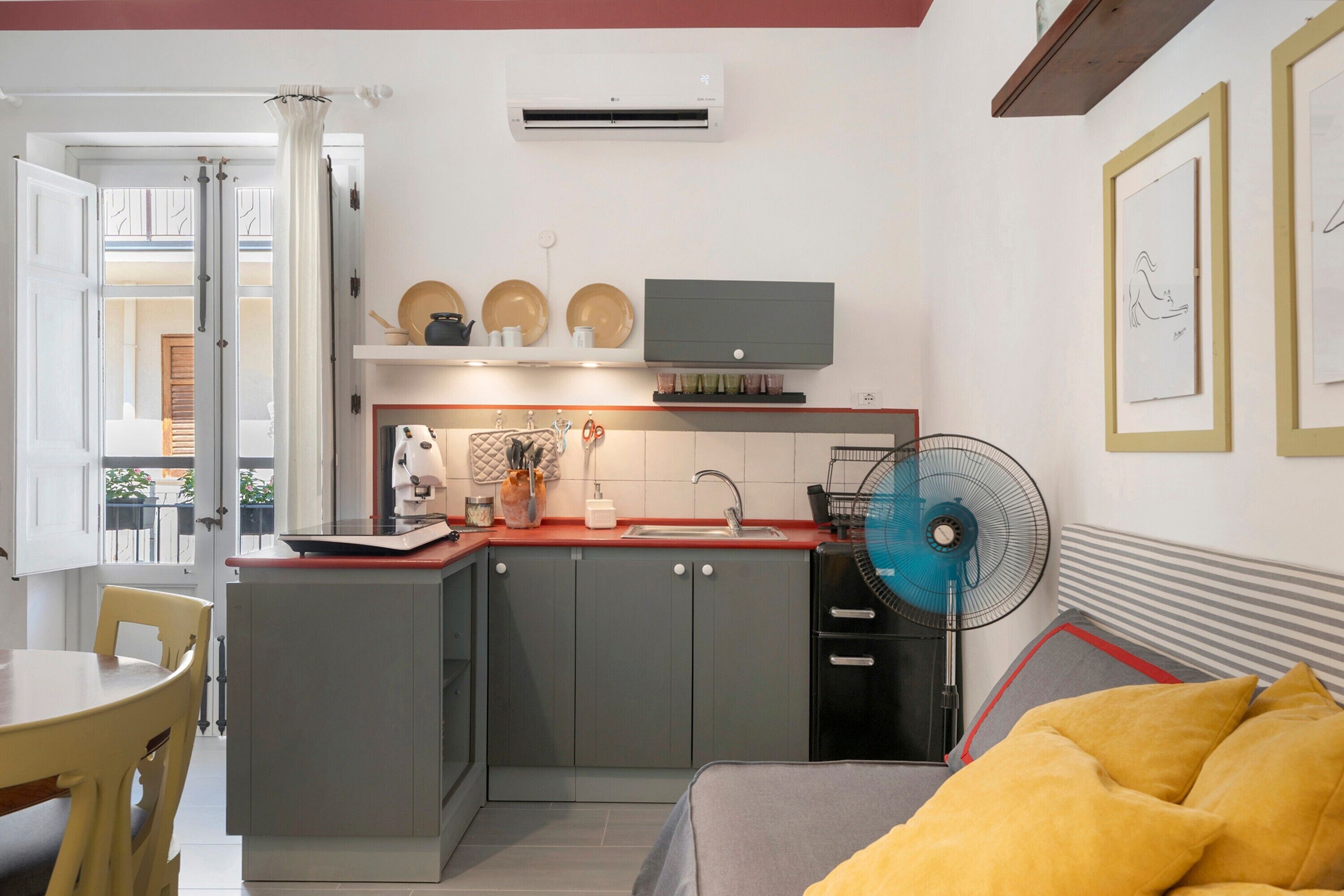 Eine kompakte Küche mit integriertem Mikrowellenherd, einer kleinen Spüle und einem Ventilator. Es gibt eine Essecke mit Tisch und Stühlen sowie ein Bett mit einem gelben Kopfkissen.