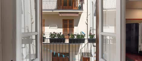 A view through a gate into a building with a balcony, a window with shutters, and potted plants.