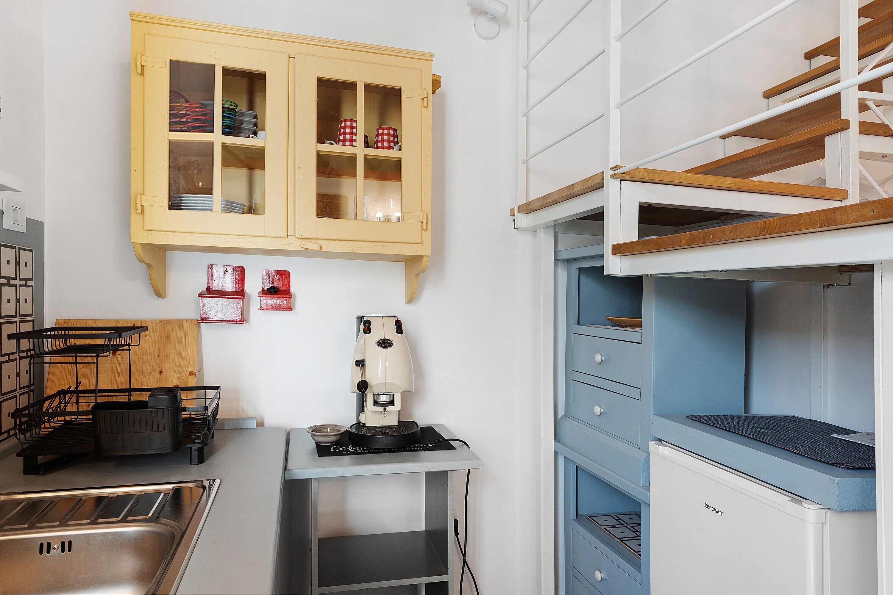 Eine kleine Küche mit Spüle, einer Kaffeemaschine, einem Schrank mit Geschirr und einer Ablage mit einer Schüssel.