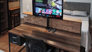 Una habitación moderna con un escritorio de madera, una silla de oficina negra, una cama con ropa de cama gris, un televisor montado mostrando 'Shrek' y cuadros en la pared.