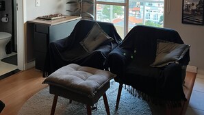 Un salón moderno con un ventanal grande, un escritorio, dos sillones y un pequeño reposapiés.