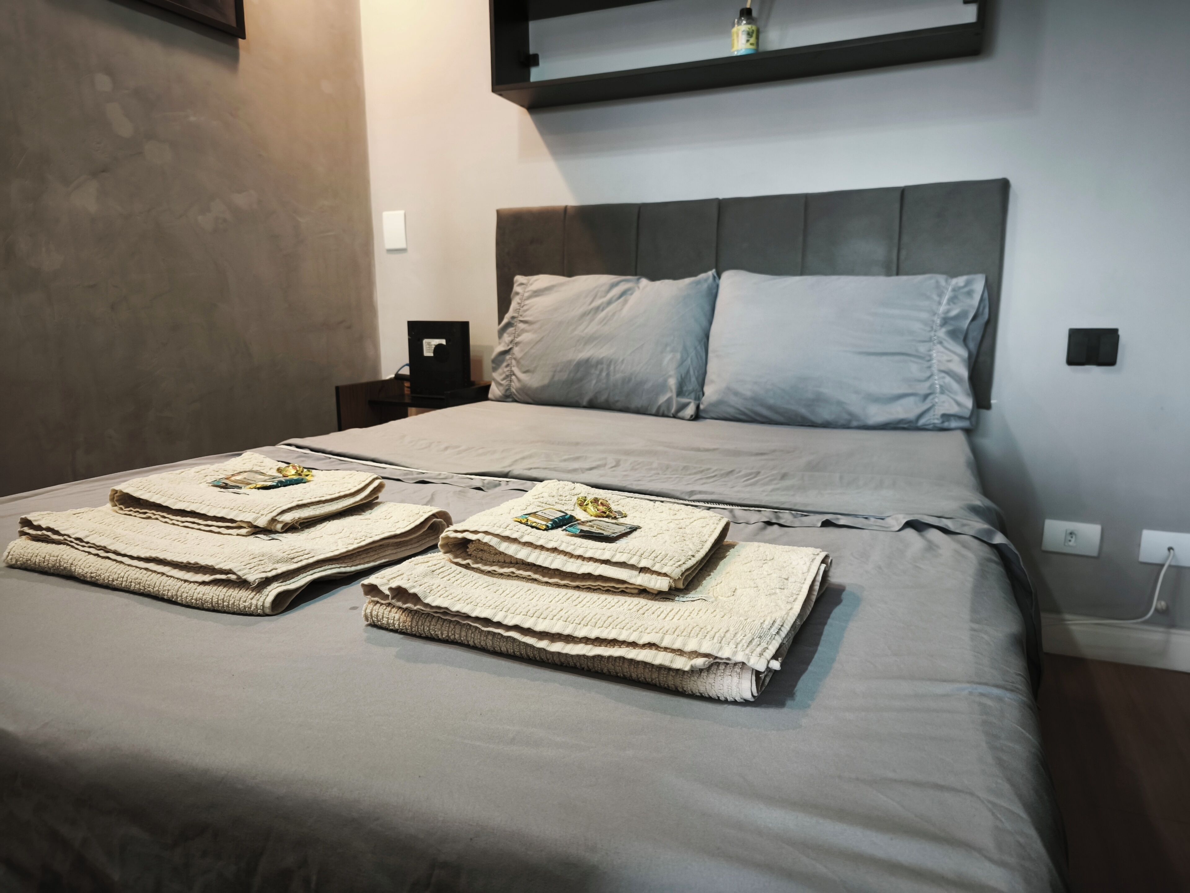 Un lit bien fait, avec une tête de lit grise et des oreillers, une table de chevet avec une lampe, et une étagère fixée au mur au-dessus du lit.