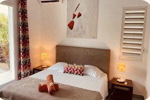 Hotellihuone, jossa on sänky, sängynpääty, yöpöytä lampulla, ikkuna, jossa on sälekaihtimet, ja seinätaideteos.