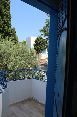 Ein Balkon mit blauer Brüstung, freier Sicht auf Bäume und Gebäude im Hintergrund sowie einem Fliesenboden.