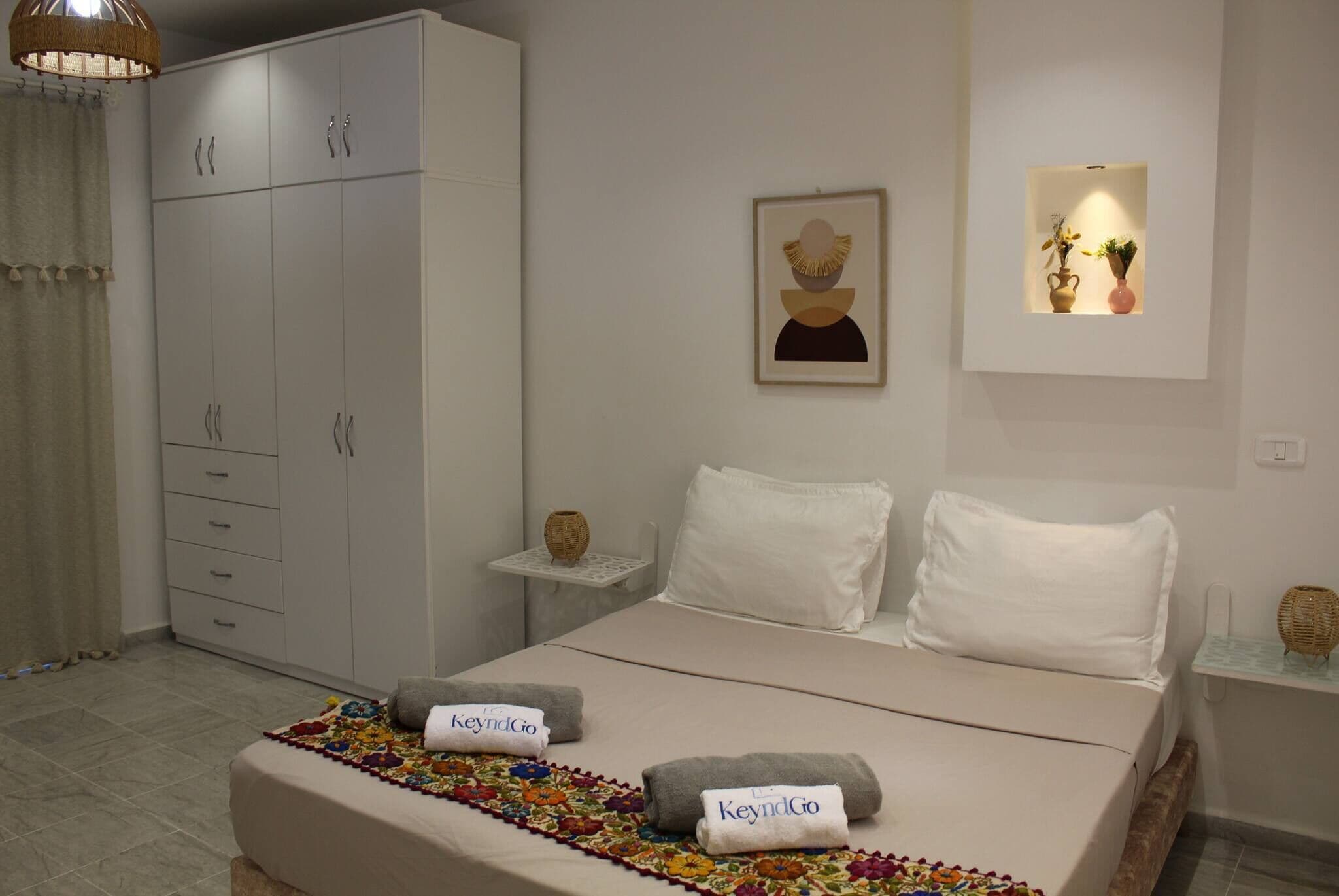 A bedroom with a bed, a wardrobe, a bedside table, a wall-mounted shelf, and a hanging light fixture.