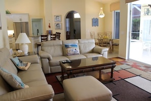 Ein Wohnzimmer mit Sofa, Hocker und Couchtisch, ein Essbereich mit Stühlen und Tisch sowie eine Küche mit Schränken und Kühlschrank.