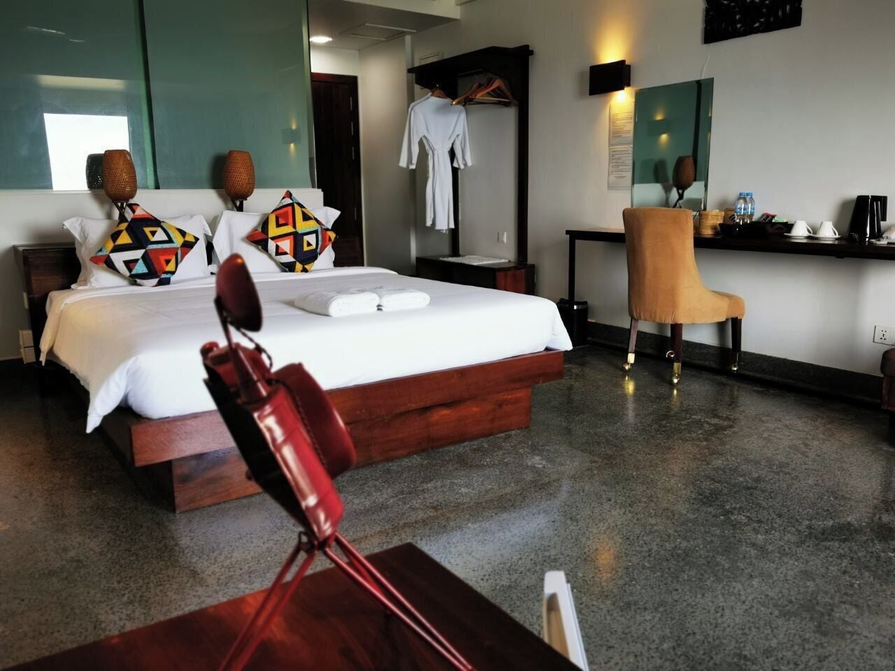 A hotel room with a large bed, a wooden side table, a chair, a desk with a chair, and a mirror.