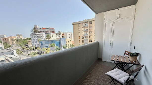 Un balcón con vistas a un paisaje urbano, una puerta blanca y una mesita con cojín y una planta.
