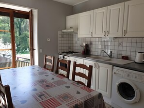 Eine Küche mit weißen Schränken, einem Esstisch mit Tischdecke, einer Waschmaschine und Blick ins Freie.