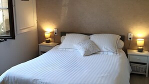 Un letto rifatto con lenzuola bianche, due abat-jour, un quadro con un gufo appeso alla parete e una finestra con una porta chiusa.