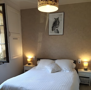 Ein ordentlich bezogenes Bett mit weißen Leinen, zwei Nachttische mit Lampen, ein gerahmtes Kunstwerk mit Eule an der Wand und ein Fenster mit geschlossener Tür.