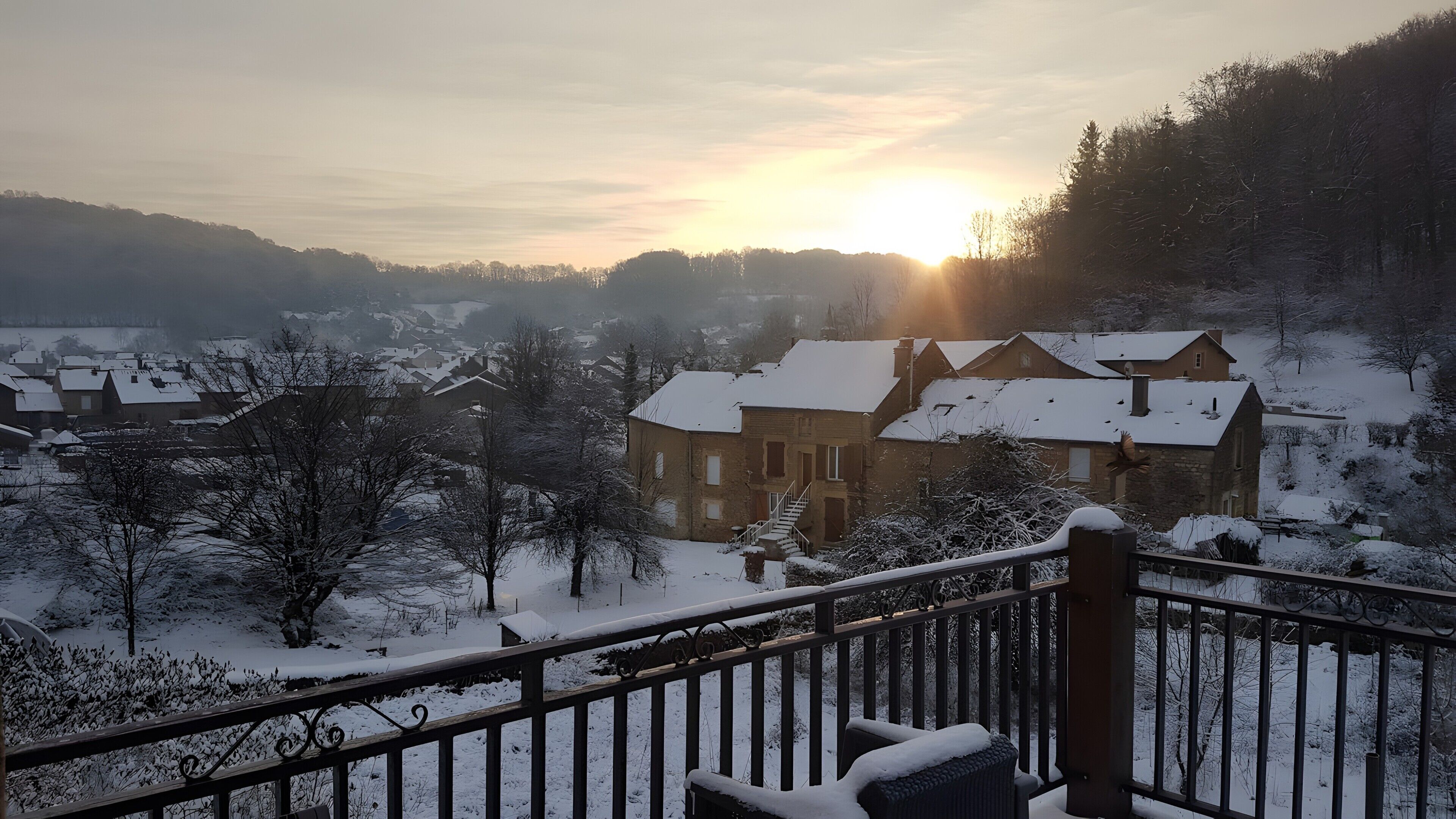 Ein verschneiter Wohnbereich mit Häusern, einem Balkon mit einem Stuhl und einem Sonnenuntergang im Hintergrund.