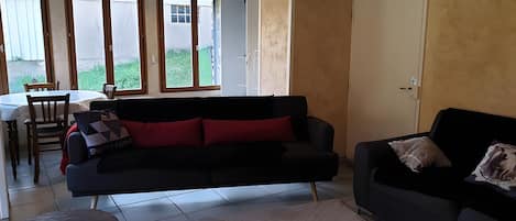 Uma sala de estar com um sofá preto, uma mesa redonda pequena e um espaço de jantar com mesa e cadeiras.