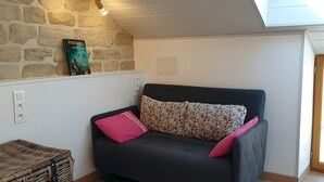 Una camera accogliente con un divano scuro, un cuscino decorato, un cesto di vimini e un muro in pietra con un libro.
