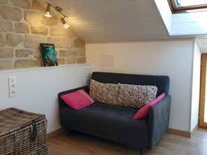 Ein gemütliches Zimmer mit einem dunklen Sofa, einem gemusterten Kissen, einem Korbwannenstuhl und einer Steinwand mit einem Buch.