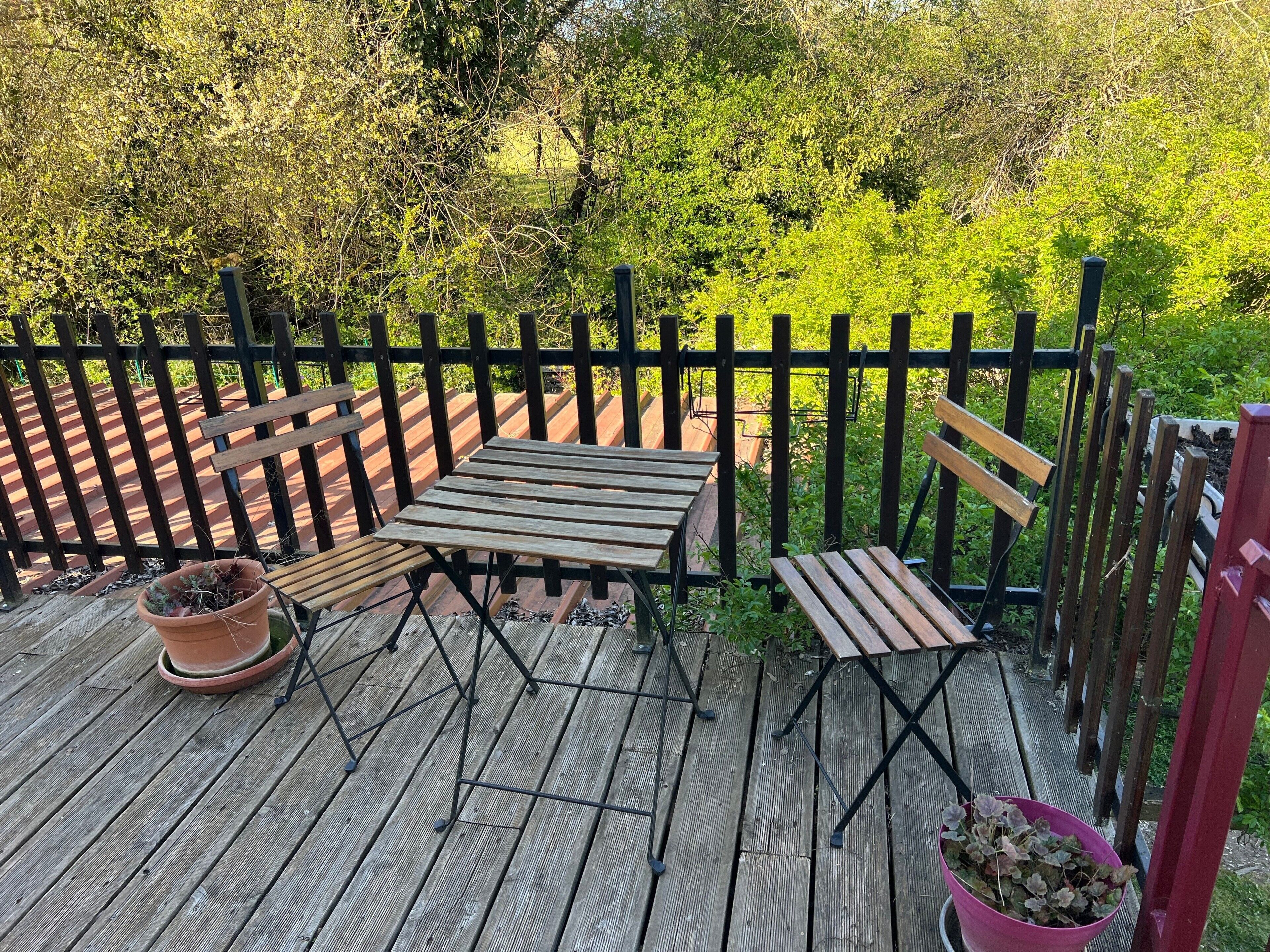 A wooden deck with a table and benches, surrounded by a black fence and lush greenery.