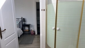 Huoneessa on sänky, yöpöytä ja pieni pöytä, jossa on lamppu. Huoneesta on ovi toiseen huoneeseen, ja siellä on lasinen suihkukaappi.