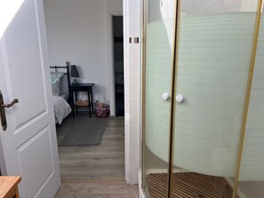 A room with a bed, bedside table, and a small table with a lamp. There is a door leading to another room and a glass shower enclosure.