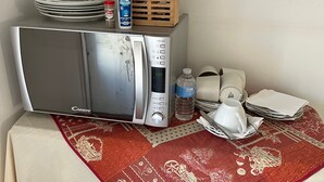 A microwave on a table with a red and white tablecloth, a stack of plates, a bottle of water, and a wooden utensil holder.