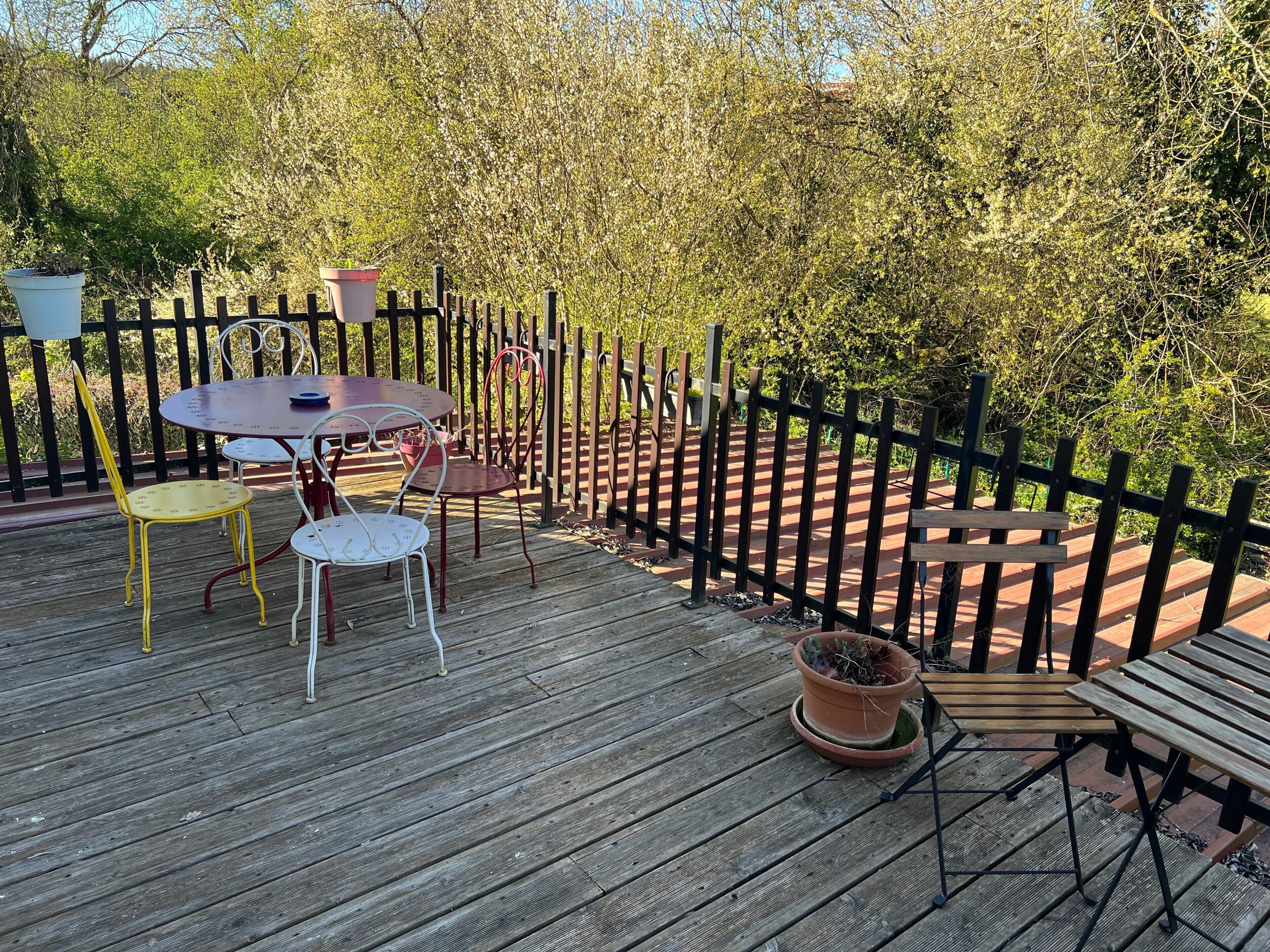 Une terrasse extérieure avec une plateforme en bois, des chaises en métal, une table ronde et un banc en bois.