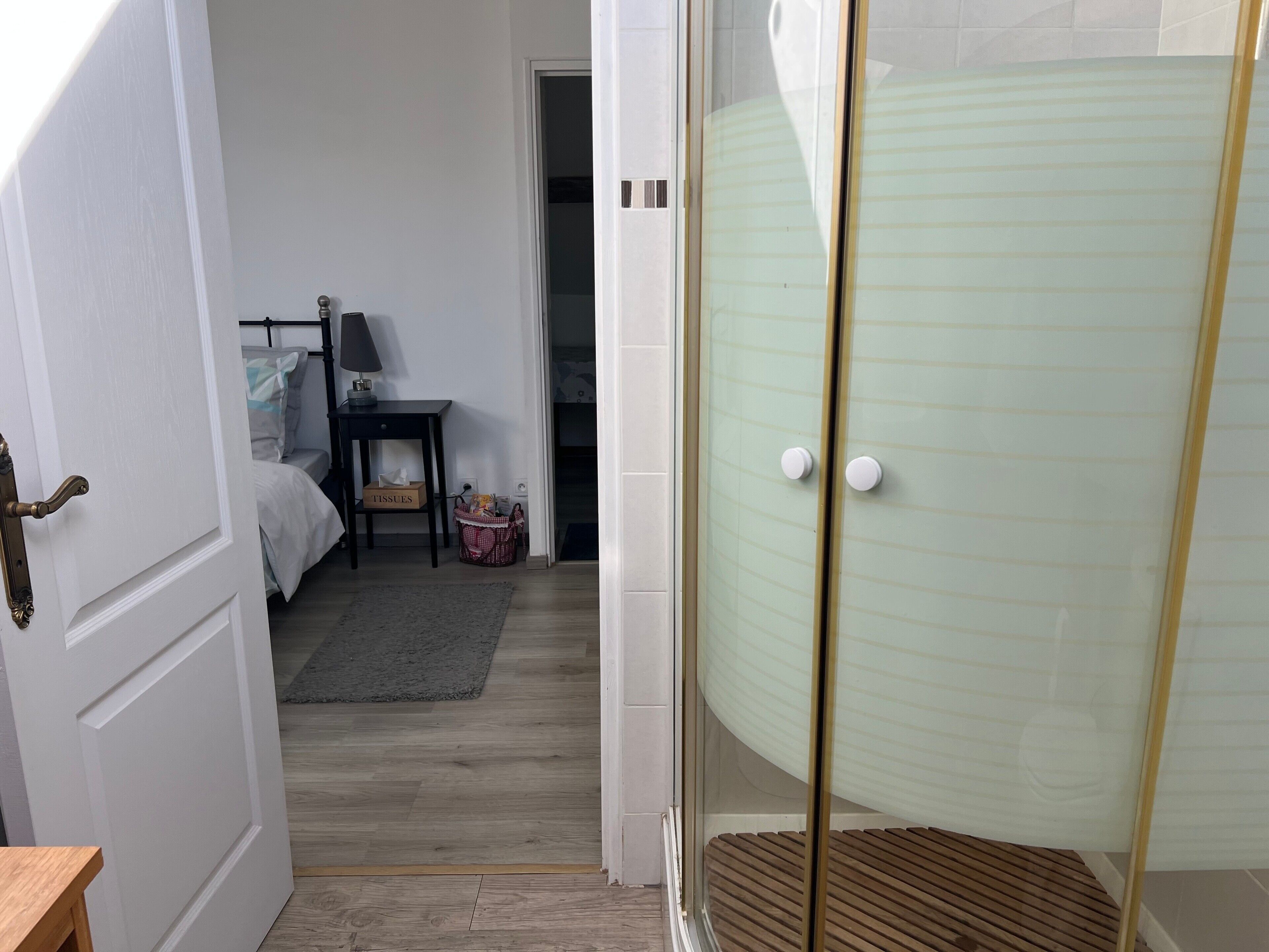 Een kamer met een bed, nachtkastje en een klein tafeltje met een lamp. Er is een deur naar een andere kamer en een glazen douchecabine.