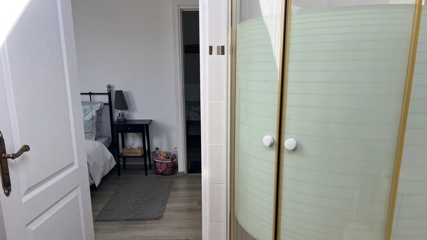 A room with a bed, bedside table, and a small table with a lamp. There is a door leading to another room and a glass shower enclosure.