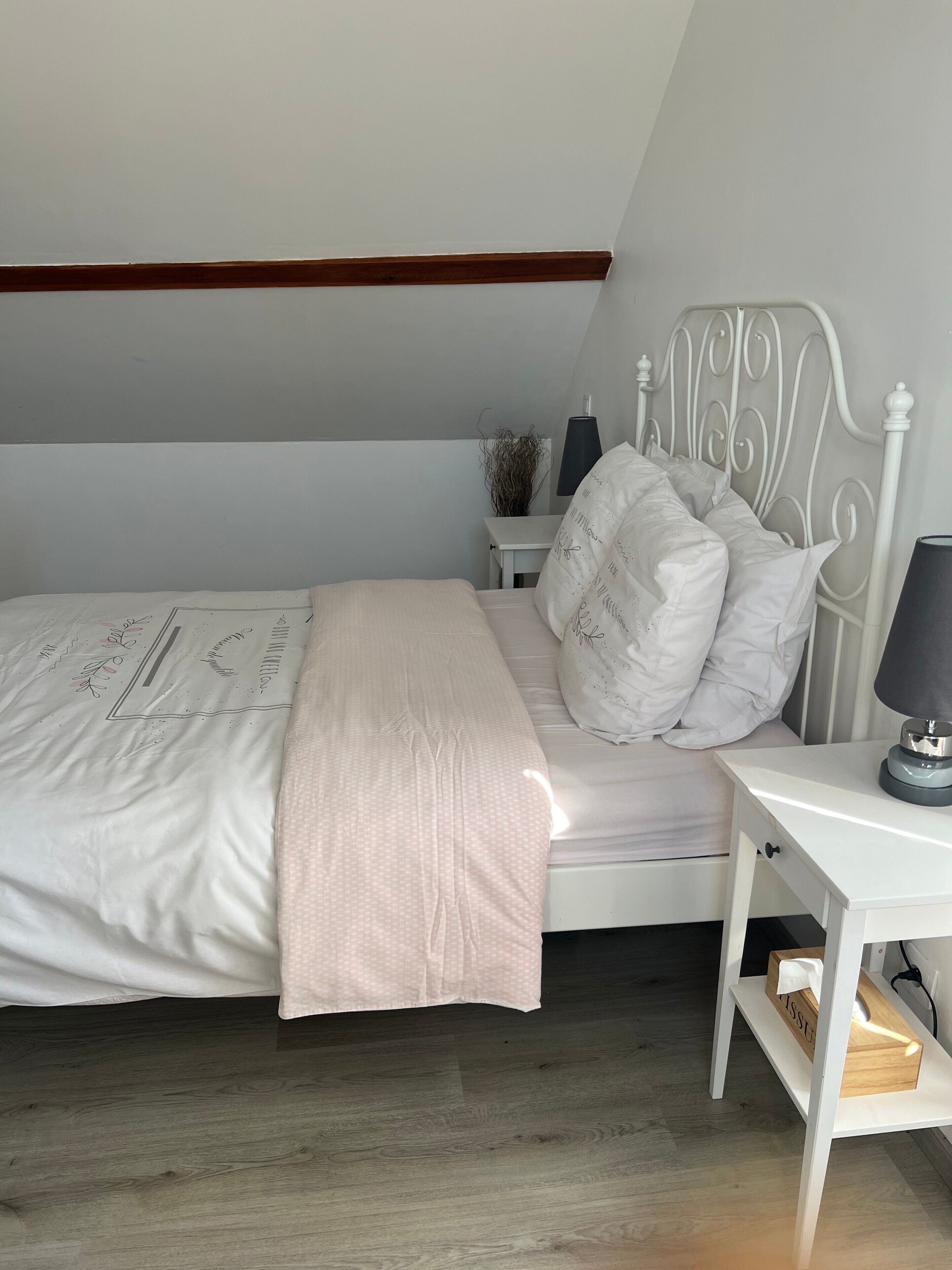 Une chambre à coucher avec un lit, une table de chevet, une lampe et une petite commode sur laquelle est posé un vase.