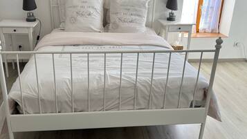 Een wit metalen bed met hoofdeinde, twee nachtkastjes met lampen, een grijs vloerkleed en een raam met gordijn.