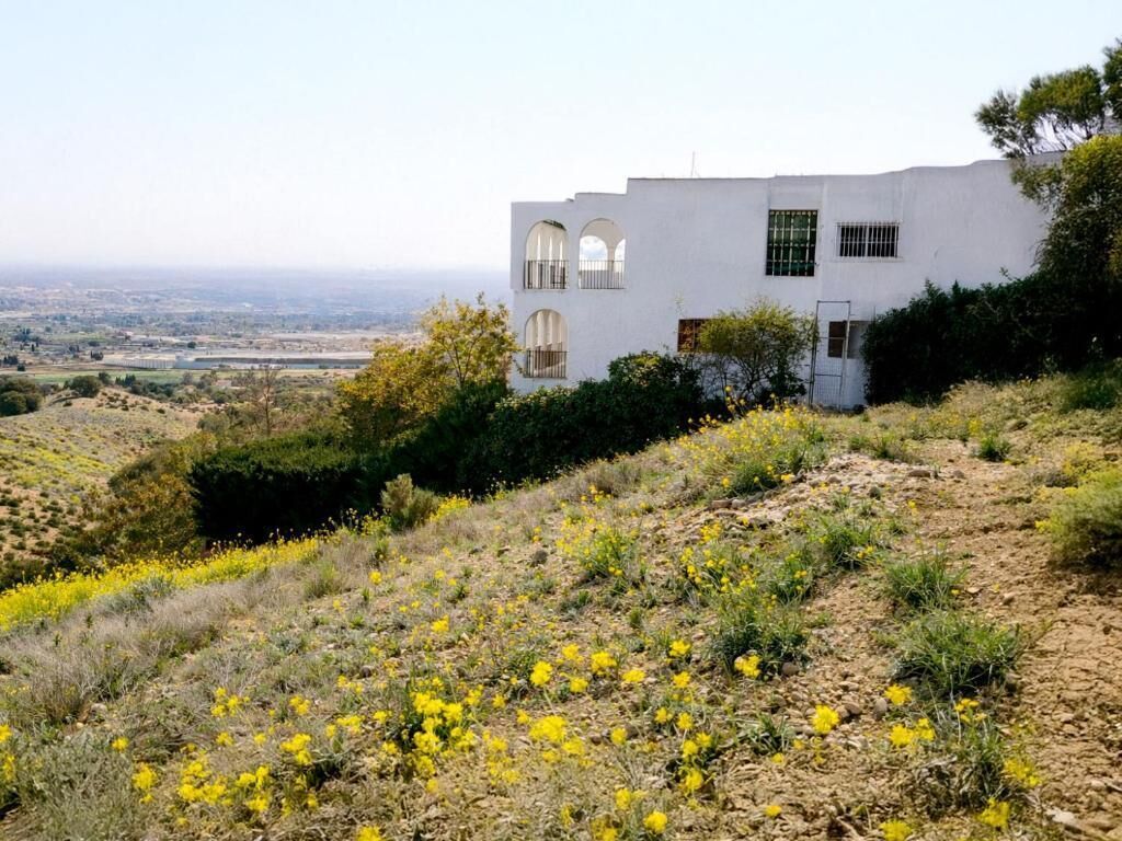 A white building with arched windows and a flat roof, surrounded by dry grass and yellow flowers.