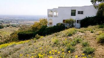 A white building with arched windows and a flat roof, surrounded by dry grass and yellow flowers.
