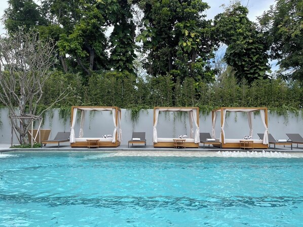 Un área junto a la piscina con cuatro camas de dosel de madera y sillones de descanso.