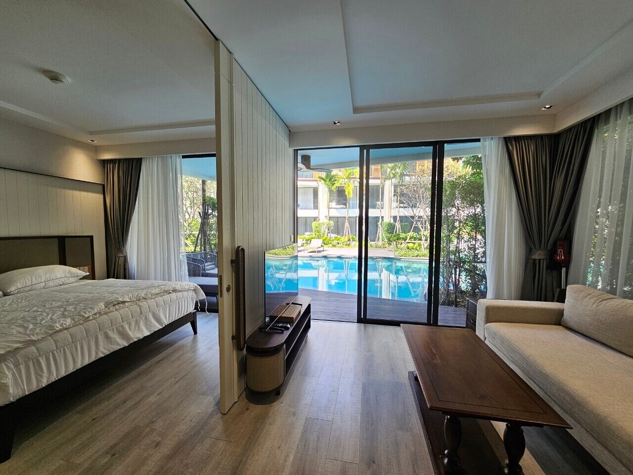 A modern hotel room with a large bed, a sofa, a small table, and a view of an outdoor swimming pool.
