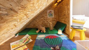 A cozy attic room with wooden walls, a green mat, a leopard plush toy, a teddy bear, a small table with books, and a hanging light fixture.