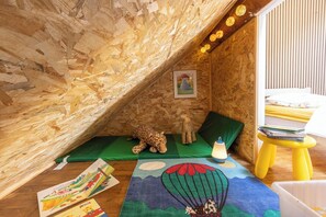 Een gezellige zolderkamer met houten wanden, een groene mat, een luipaardknuffel, een teddybeer, een klein tafeltje met boeken en een hangende lamp.