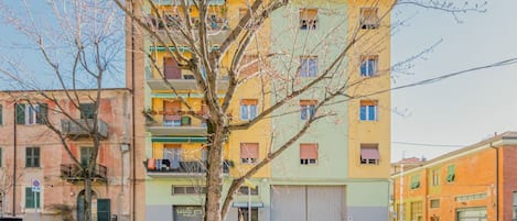 Monikerroksinen rakennus, jossa on parvekkeita, etualalla puu ja parkissa olevia autoja.