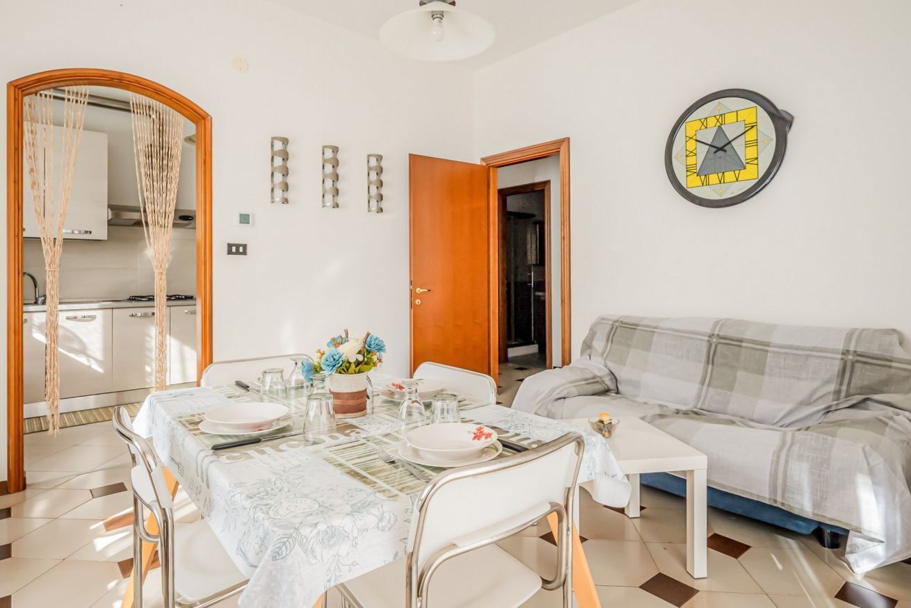 A dining area with a table set for four, a sofa, a clock, and a kitchen visible through an open door.
