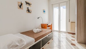 Yhden hengen sänky, jossa on valkoinen ja ruskea päiväpeite, työpöytä tuolilla, lamppu ja ikkuna, josta on näkymä.