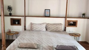 Una cama con dosel de madera, con un edredón blanco y dos almohadas. Hay dos mesitas de noche con lámparas, un cuadro enmarcado en la pared y una pequeña planta en maceta a la izquierda.