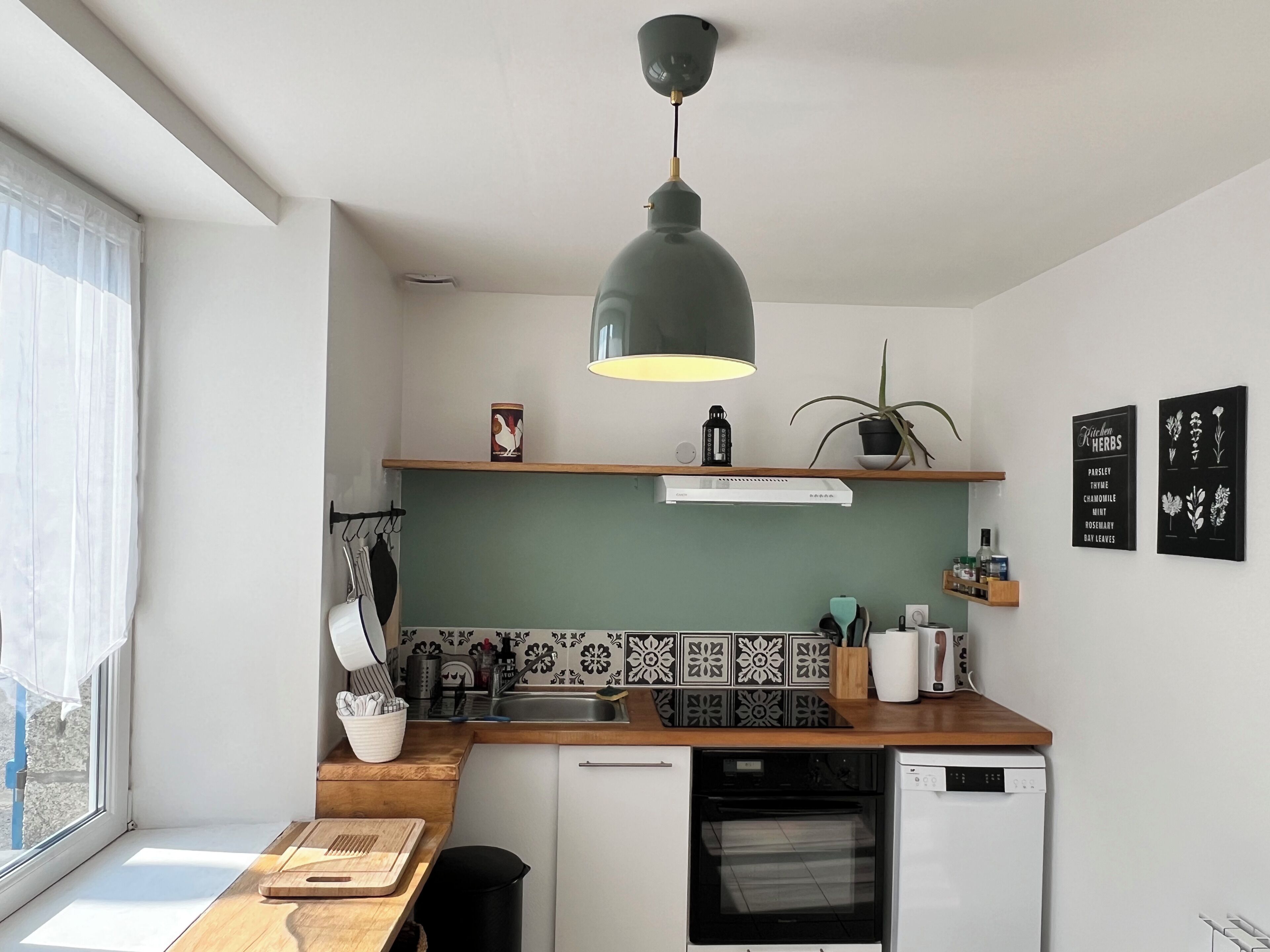 A modern kitchen with a built-in oven, a dishwasher, a hanging light fixture, and a window with sheer curtains.