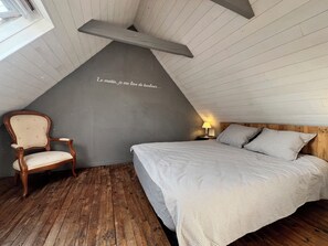 Une chambre avec un plafond en pente, une chaise en bois, un lit avec une literie à rayures et un mur orné d’une phrase en français.